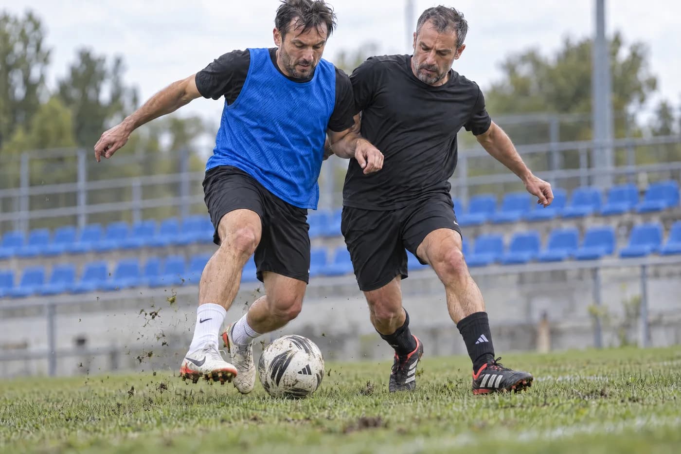 שני שחקנים בשחור נאבקים על הכדור באצטדיון, אחד עם גופיית אימון כחולה, יציע כחול ברקע